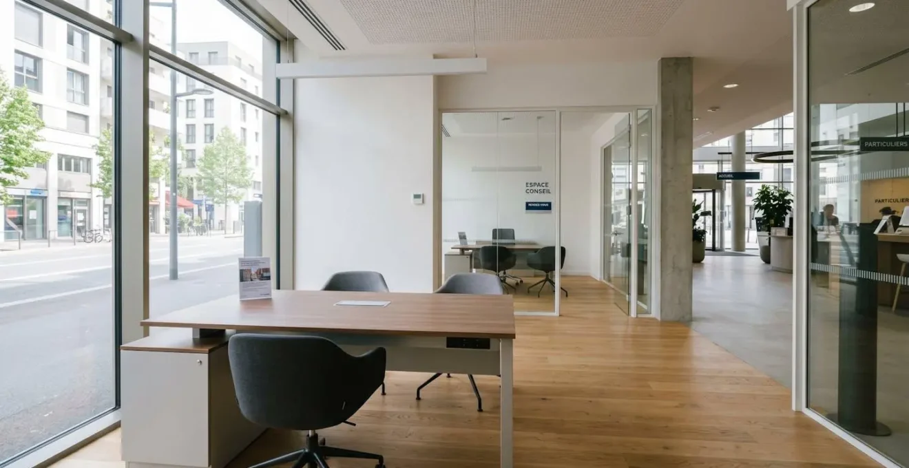 Vue grand angle d'un espace de conseil bancaire contemporain avec bureau épuré, chaises design et larges fenêtres laissant entrer une lumière naturelle abondante, sans présence humaine au premier plan